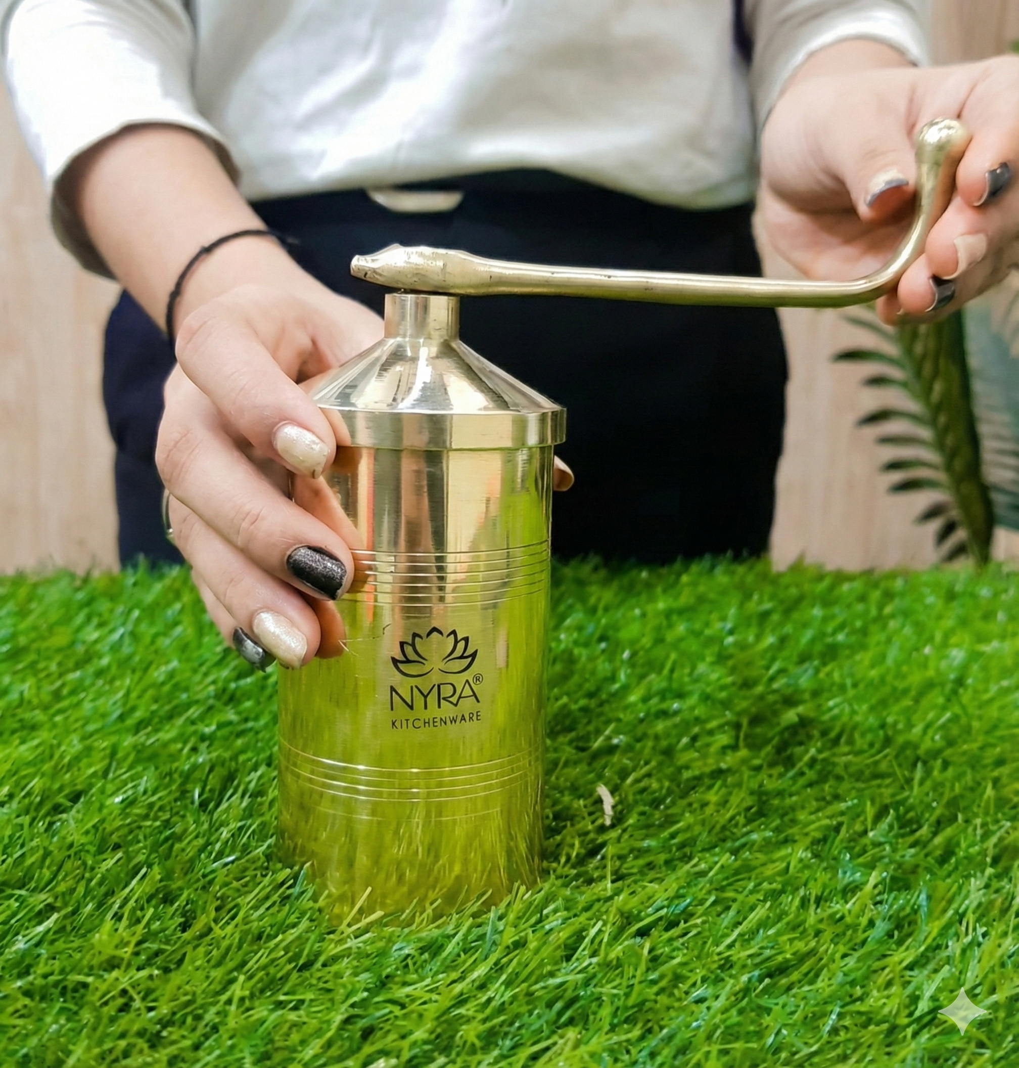 Person using a gold manual juicer with 'Nyra Kitchenware' branding on a grassy surface.